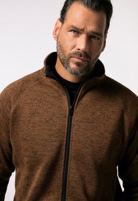 Giacca zip-up marrone con texture, colletto e dettagli neri. Caratterizzata da una cerniera frontale completa e maniche raglan per facilitare i movimenti.