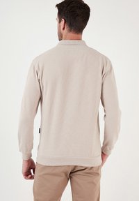 Világos bézs hosszú ujjú kötött póló, bordázott textúrával, hajtott gallérral és fitted hem-mel. Minimalista dizájn, látható logó nélkül.
