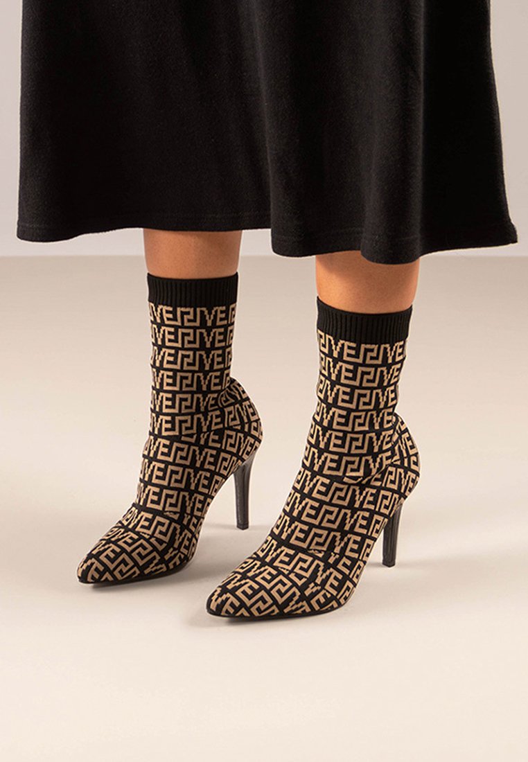 Botines de calcetín en negro y beige con un alto tacón de aguja, que presentan un patrón geométrico en toda la tela y un puño de tobillo acanalado.