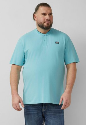 Mężczyzna z brodą i związanymi włosami, ubrany w jasnoniebieską koszulkę polo z krótkim rękawem i ciemnoniebieskie dżinsy, stojący na jednolitym tle.