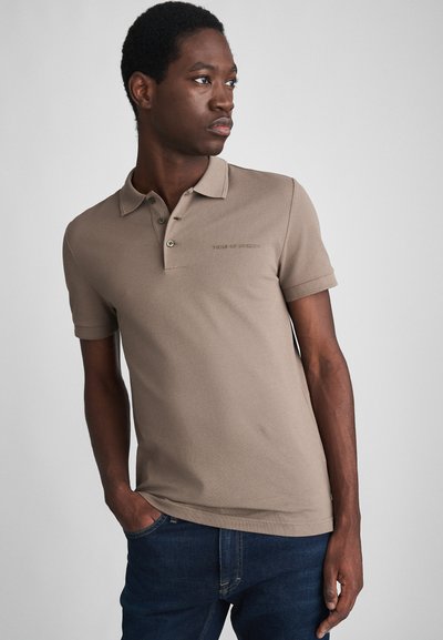 Beige színű polo felső, puha tapintású, klasszikus gallérral, három gombbal és diszkrét márkaboltítással a mellen. Kék farmernadrággal párosítva.