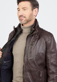 Bruine leren jack met ritssluiting, textuurafwerking, twee ritszakken en een staande kraag. De binnenvoering heeft een gefigureerde stof.
