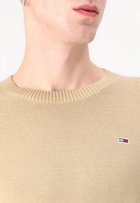 Beige strikkegenser med ribbet rund hals. Har et lite logomerke i rødt, hvitt og blått på venstre brystområde.