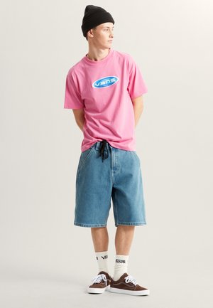 Różowa koszulka z krótkim rękawem z logo "Vans", zestawiona z luźnymi szortami dżinsowymi, białymi skarpetkami i brązowymi trampkami, dopełniona czarną czapką beanie.
