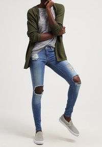 Musta vihreä neuletakki, harmaa raidallinen t-paita, kuluneet siniset farkut, joissa on polvihaavoja, ja harmaat liukuvat kengät. Rentoa kerrospukeutumista.