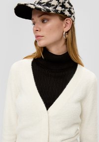 Cremfarbener Cardigan über einem schwarzen, gerippten Rollkragenpullover, mit V-Ausschnitt, Frontknöpfen und einer gemusterten schwarz-weißen Mütze.