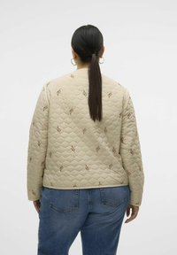 Gequilte beige jas met bruine geborduurde accenten. Heeft een ontspannen pasvorm, lange mouwen en een gebande zoom, gecombineerd met denim jeans.