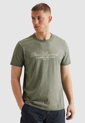 Mann trägt grünes "Pmc Legend Cargo Goods Since 1958" T-Shirt und dunkle Hose, schaut nach links, Hände teilweise in den Taschen.