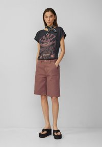 Grafische t-shirt in donkergrijs met een bedrukt ontwerp, gecombineerd met licht mauve korte broek. Zwarte platform sandalen maken de outfit af.