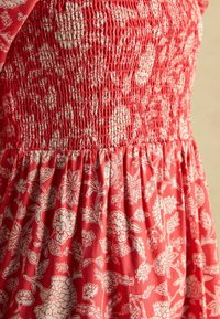 Rotes Blumenmusterkleid mit weißen, filigranen Mustern. Verfügt über ein gerafftes Oberteil und einen weit schwingenden Rock. Weicher, strukturierter Baumwollstoff.