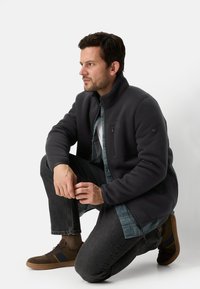 Donkergrijs fleecejack met een hoge kraag en een zak met rits, gedragen over een geruit overhemd met donkergrijze spijkerbroek en bruine schoenen.