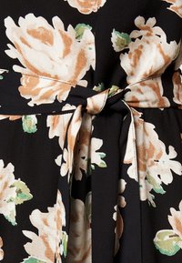 Textilie s květinovým vzorem v černé barvě, s velkými béžovými a zelenými květinami. Zahrnuje látkový opasek uvázaný v pase, s hladkou strukturou.