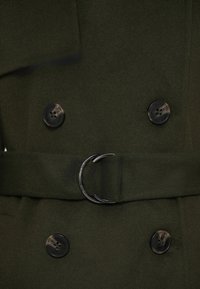 Olijfgroen wollen jas met een dubbelgeknoopt ontwerp, voorzien van twee zwarte knopen en een metalen riemgesp. Zachte textuur en op maat gemaakte pasvorm.