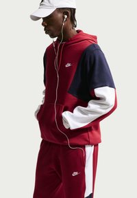 Jeune personne portant un survêtement Nike rouge, bleu marine et blanc, les mains dans les poches, casquette blanche et écouteurs, regardant vers le bas.