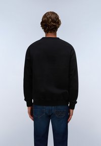 Uomo con capelli castani ricci che indossa una felpa nera e jeans blu, visto di spalle contro uno sfondo grigio chiaro.