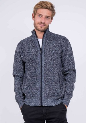 Homme aux cheveux châtain clair porte un pull zippé gris texturé et un pantalon noir, debout les mains dans les poches devant un fond uni.