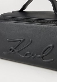 Mustal tekstuuriga sünteetiline kott, millel on silmapaistev reljeefne "Karl" logo, lukuga sulgemine ja väike sang üleval.