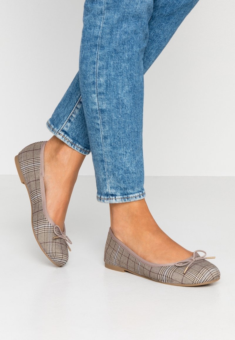 Pieds portant des ballerines à carreaux beige avec petits nœuds, associées à un jean bleu roulé sur un fond clair.