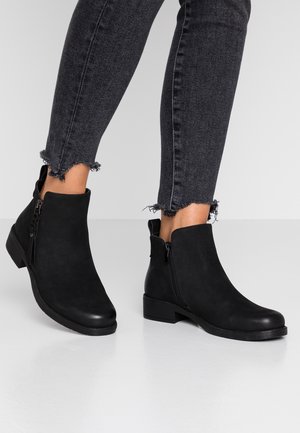 Bottines noires en suedine, dotées d'une fermeture éclair sur le côté et d'un petit talon carré. Portées avec un jean noir effiloché.