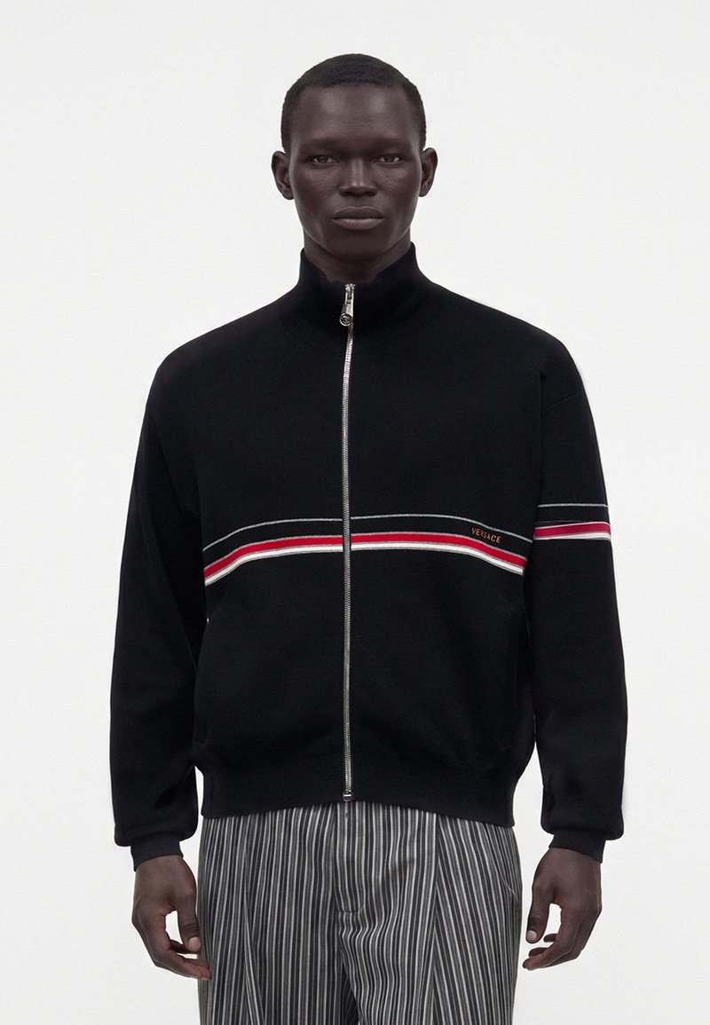 Homme portant une veste noire zippée avec des rayures horizontales rouges, blanches et grises et un petit logo Versace sur la poitrine, assortie à un pantalon gris rayé.
