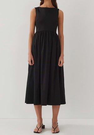 Nainen, joka pukeutunut hiukseton mustaan midimekkoon, jossa on vartalonmyötäinen päälliosa ja kerrostettu hame, yhdistettynä mustiin korkokenkiin.