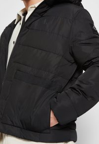 Giubbotto imbottito nero con cappuccio, caratterizzato da una chiusura zip, tasche laterali e un tessuto testurizzato con cuciture orizzontali. Accentuato da loghi.