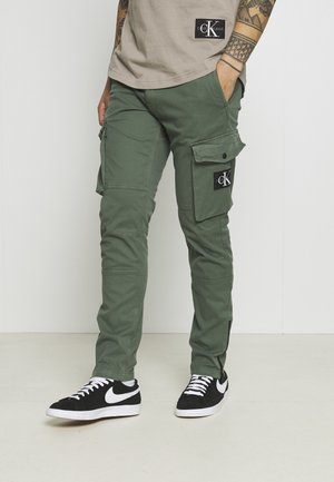 Osoba w oliwkowych spodniach cargo z dużymi kieszeniami, beżowej koszuli Calvin Klein, czarnych sneakersach Nike i z wytatuowaną ręką, stojąca w pomieszczeniu.