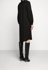 Schwarzes, langärmeliges Kleid mit lockerer Passform, das ausgestellte Ärmel und einen asymmetrischen Saum hat. Kombiniert mit schwarzen knielangen, ledernen Stiefeln mit Absatz.