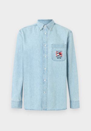 Camisa de mezclilla azul claro de manga larga con botones y bolsillo en el pecho que presenta un parche con el logo de Tommy Hilfiger en rojo, blanco y azul.