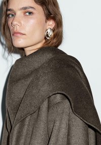 Cappotto marrone, strutturato e over size con colletto alto e design drappeggiato; abbinato a un grande orecchino d'argento curvo.