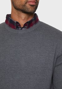 Maglione grigio a coste con scollo rotondo indossato sopra una camicia a quadri rossa e blu, con il colletto a scacchi visibile sotto.