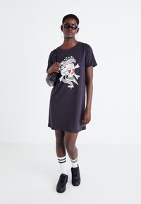 Vestido corto negro con un gráfico frontal que presenta una calavera y huesos. Se lleva con zapatillas negras y calcetines blancos con rayas negras.