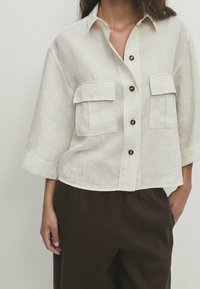Light beige, short-sleeve button-up shirt with two chest pockets, brown buttons, and a relaxed fit, made from a textured fabric.