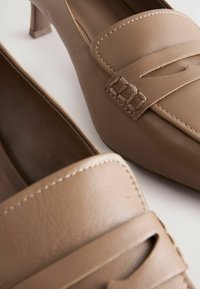 Braune Lederloafers mit niedrigem Absatz, die ein gesticktes Design und einen geflochtenen Akzent über dem Spann aufweisen. Glatte Textur und klare Linien.