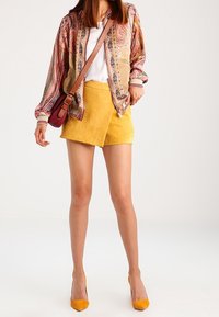 Färgglad mönstrad bomberjacka med beige bas, gula mockashorts, gula klackar och en liten vinröd axelväska.