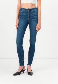 Jeans albaștri cu talie înaltă, cu o croială slim, din denim. Au cinci buzunare, fermoar și detalii de cusătură minime. Asortați cu tocuri cu slingback negre.