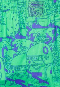 Shorts colorate cu un fundal verde și ilustrații de personaje cartoon în diverse scene, și o aplicație decorativă pe talie.