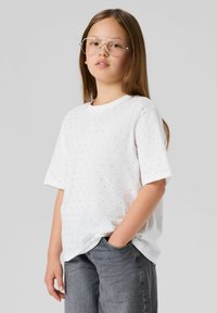 Biała koszulka z krótkim rękawem o teksturowanym wzorze małych kropek, zestawiona z szarymi dżinsami. Modelka ma długie włosy i nosi okulary.