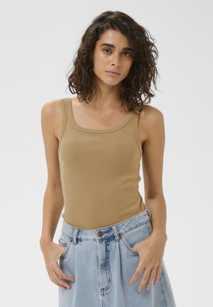 Femme aux cheveux bouclés et foncés, portant un haut beige sans manches et un jean taille haute bleu clair, debout devant un fond uni clair.