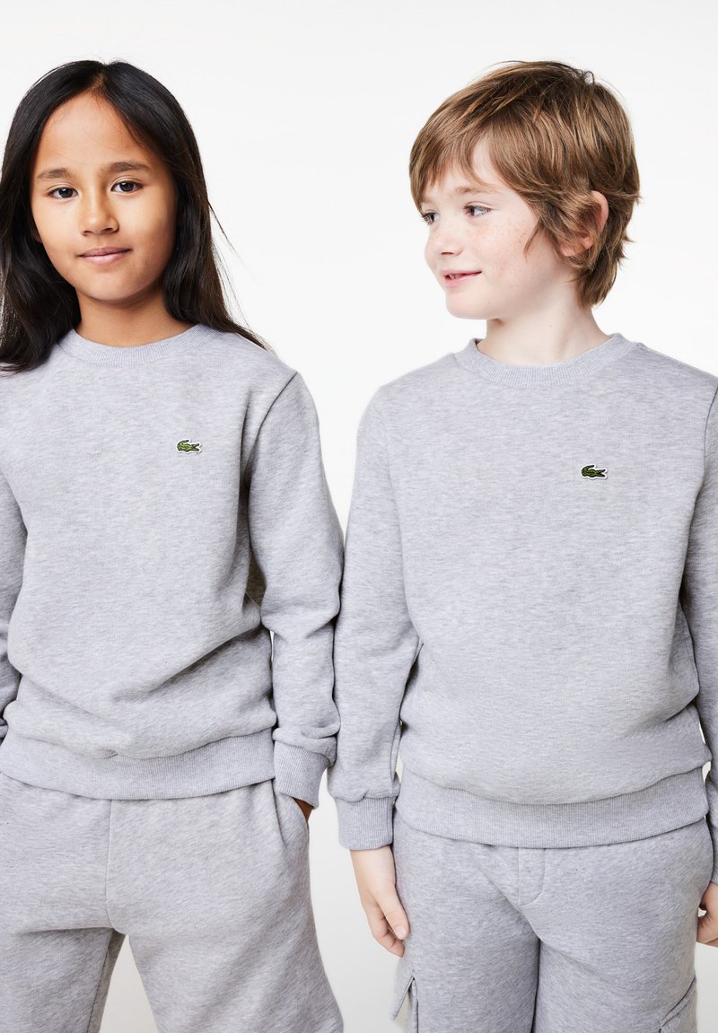 Dvoje djece nose uskla�đene sive dukserice s rebrastim rukavima i pojasom. Svaki ima mali zeleni Lacoste logo na grudima.