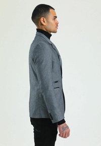 Grau maßgeschneiderter Blazer, strukturierter Stoff, einreihiges Design, schwarze Akzente, zwei Fronttaschen, Slim Fit, getragen über einem schwarzen Rollkragenpullover.