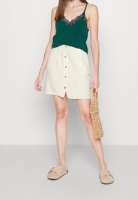 Canotta verde con pizzo, gonna crema con bottoni davanti, scarpe beige senza lacci e borsa intrecciata. Tessuto morbido con design casual.