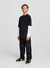 Czarna i biała koszula z długim rękawem o luźnym kroju, zestawiona z czarnymi spodniami typu cargo z białym przeszyciem i wieloma kieszeniami. Czarne trampki.