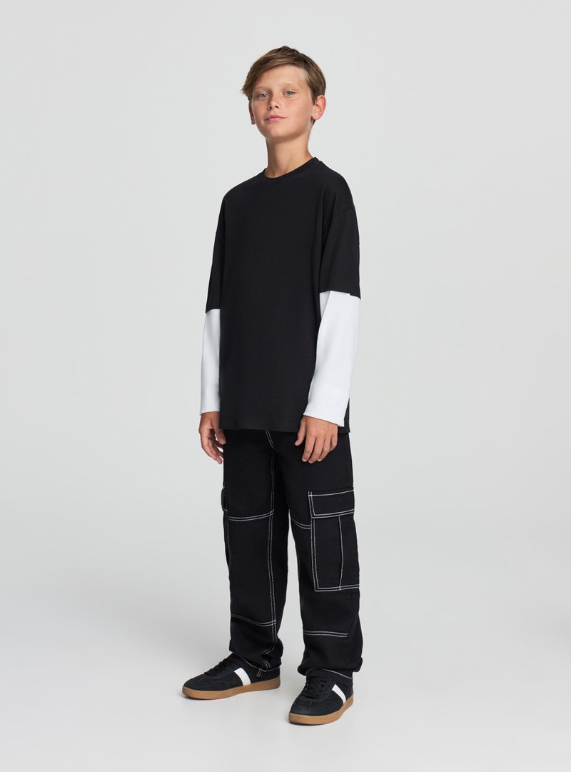 Czarna i biała koszula z długim rękawem o luźnym kroju, zestawiona z czarnymi spodniami typu cargo z białym przeszyciem i wieloma kieszeniami. Czarne trampki.