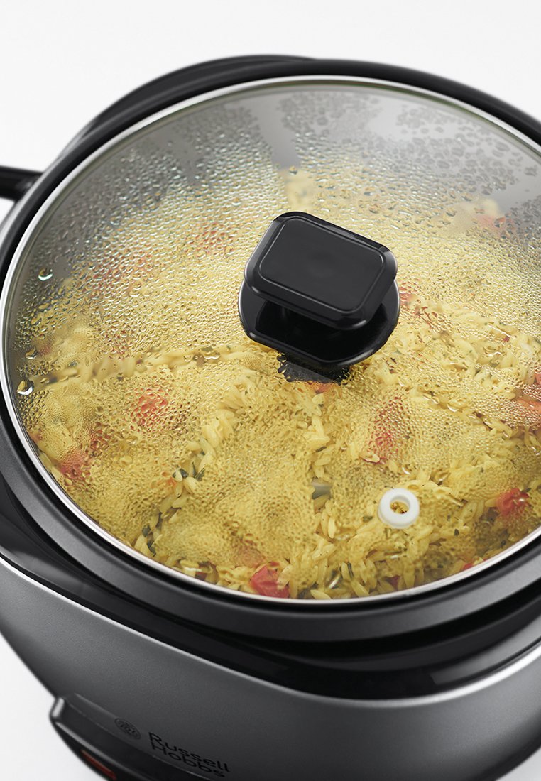 Olla arrocera negra con tapa de vidrio transparente, visible la condensación del vapor. Contiene arroz cocido mezclado con hierbas y verduras.