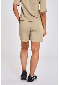 Person klädd i beige shorts och matchande topp, stående med händerna bakom ryggen, iförd svarta och vita sneakers mot en vit bakgrund.