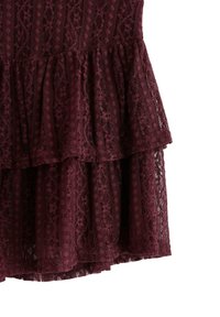 Bordeaux kantstof met gelaagde ruffles, featuring ingewikkelde patronen en een semi-doorzichtige textuur op een witte achtergrond.