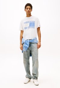 Camiseta blanca con diseño gráfico azul, combinada con jeans de un azul claro. Una camisa de patrón azul está atada a la cintura. Zapatillas blancas.
