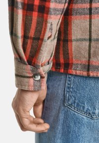 Flanellhemd mit rot, schwarz und grün kariertem Muster. Verfügt über einen Knopf-Manschette und wird mit blauen Jeans kombiniert. Strukturiert.