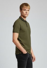 Olivově zelené polo tričko s krátkými rukávy a černým límcem s vzorem, s tříknoflíkovou légou a bočními rozparky pro lepší pohyb.